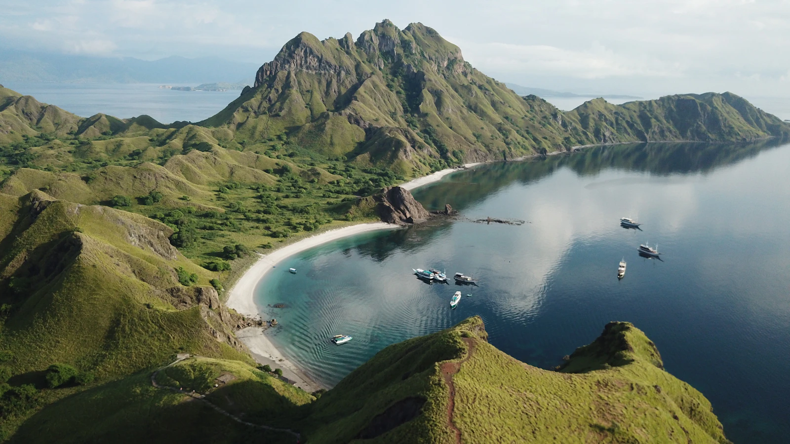Aerial view of a green ridged island above a bay with a pale curved beach and moored boats